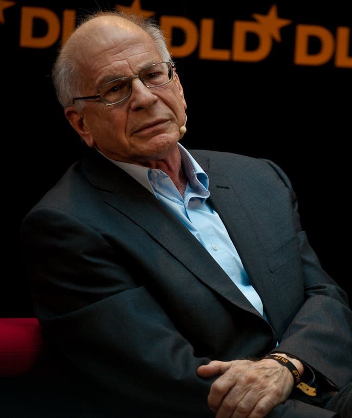 A man, wearing glasses and a suit, sits with his arms crossed. He has a microphone on his cheek. The background features repeated "DLD*" text.