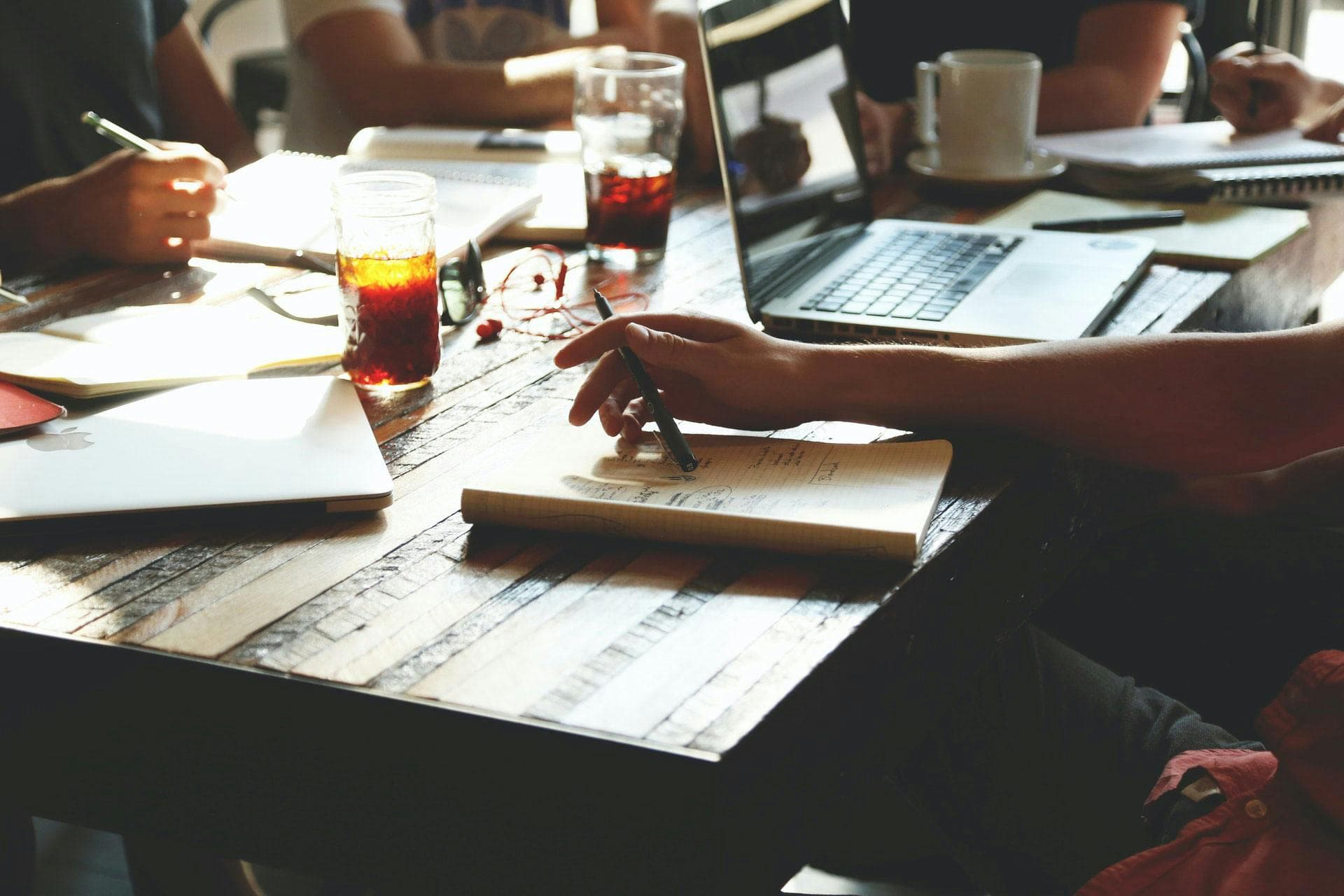 People taking notes and using laptops in a meeting, with drinks and other office items spread across a wooden table. Sunlight filters through, creating a collaborative, relaxed environment.