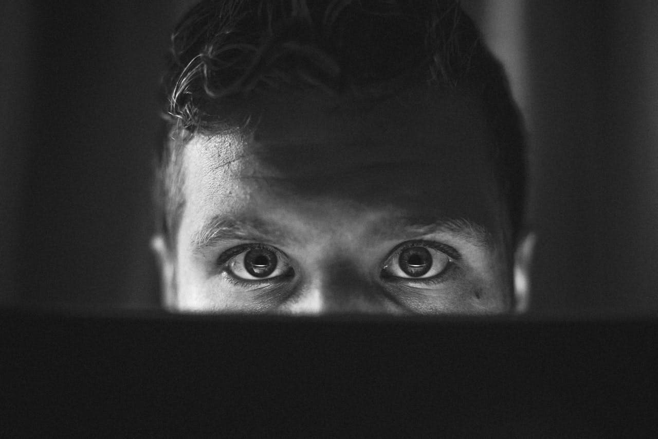 A person is staring intently at a screen in front of them, with their face partially illuminated and surrounded by darkness in a dimly lit room.