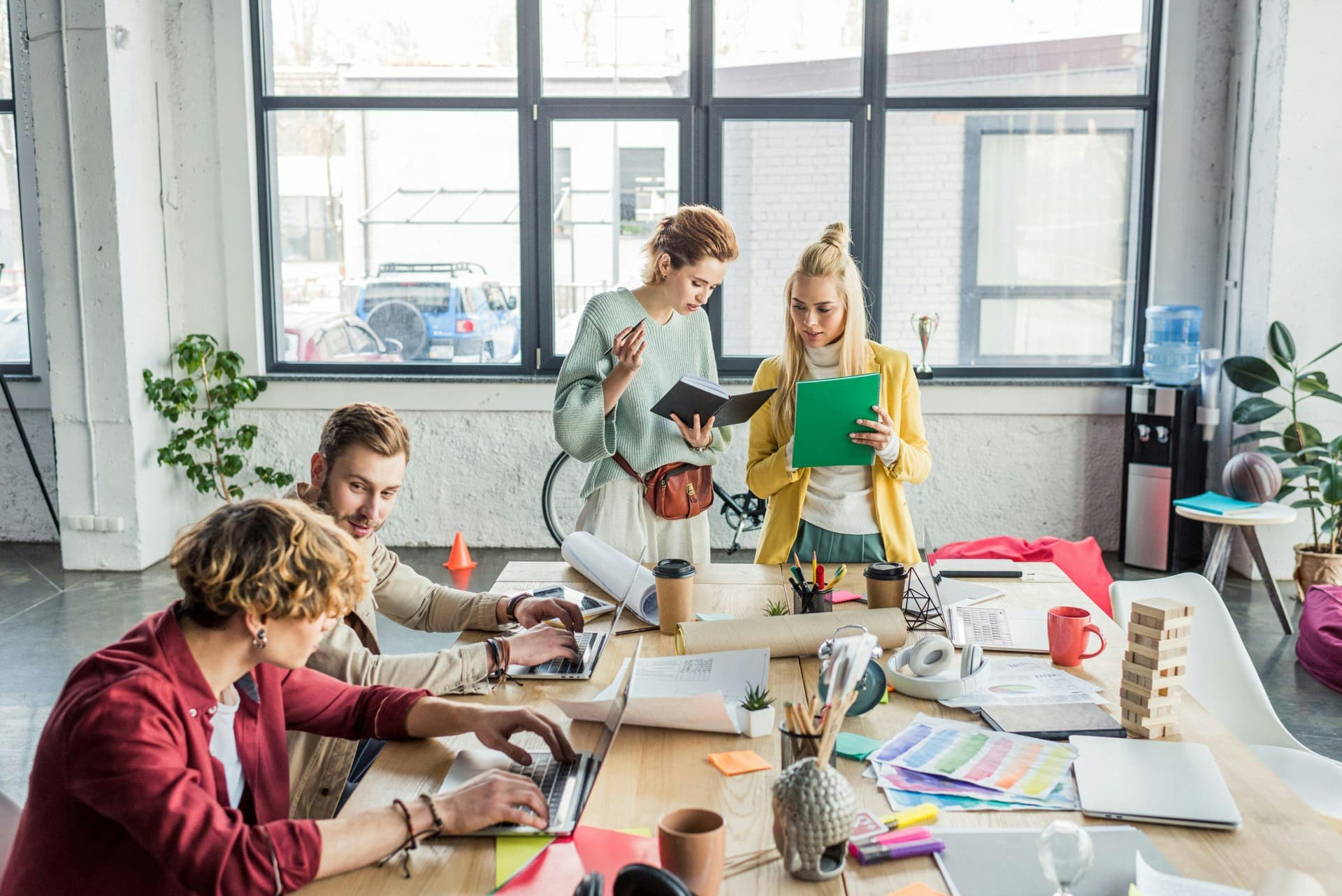 People discussing and working on laptops around a large table in a modern office with big windows, plants, and various office supplies, including notebooks, coffee cups, and sketches.