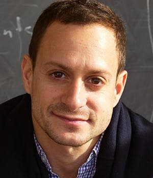 A person with short hair and a slight smile is looking directly at the camera, with a chalkboard featuring partially visible writing in the background.