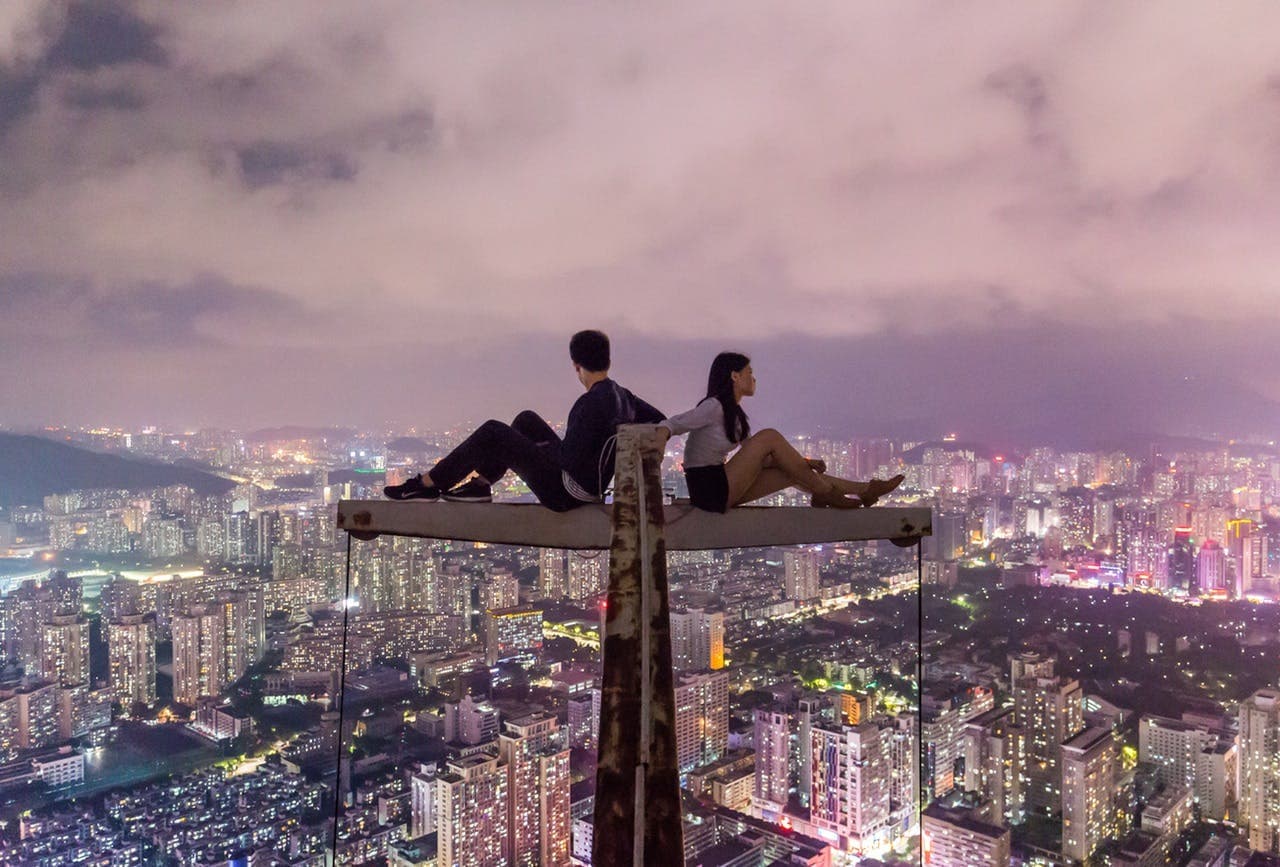 Two people sit back-to-back on a narrow beam, high above a brightly-lit, sprawling cityscape at night, with buildings and lights extending far into the distance.
