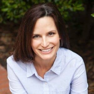 A woman with shoulder-length brown hair smiles while sitting outdoors in a natural setting, with greenery behind her and brick ground partially visible. She wears a light blue button-up shirt.