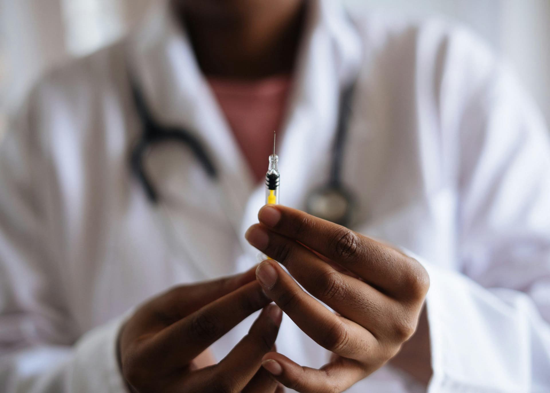 A person in a white lab coat holds a syringe with an amber liquid, readying it for use, in what appears to be a medical setting.