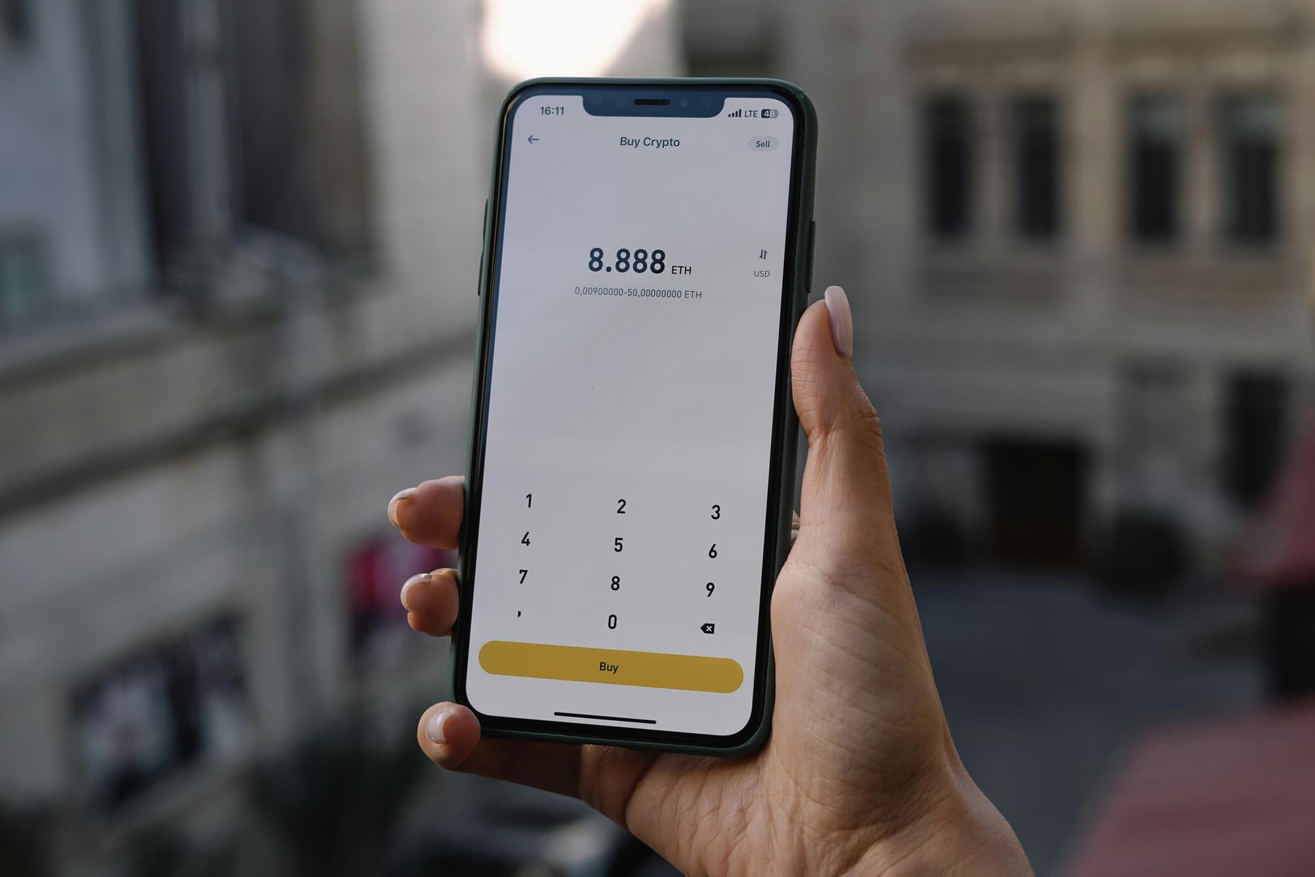 A hand holds a smartphone displaying a cryptocurrency purchase screen showing 8.888 ETH, with "Buy" and numeric keypad below. Background shows blurred urban buildings.