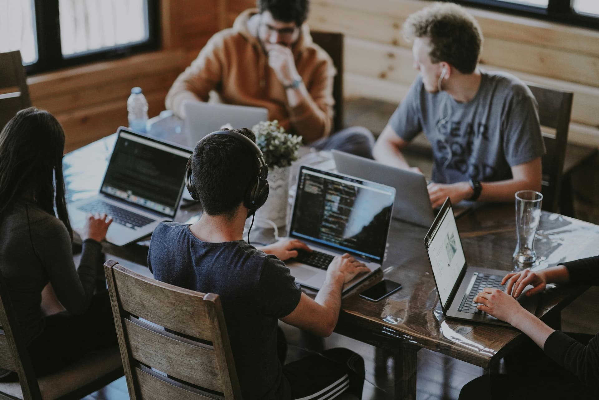 Five people are sitting around a wooden table, working on laptops in a casual, well-lit room with wooden walls and large windows.