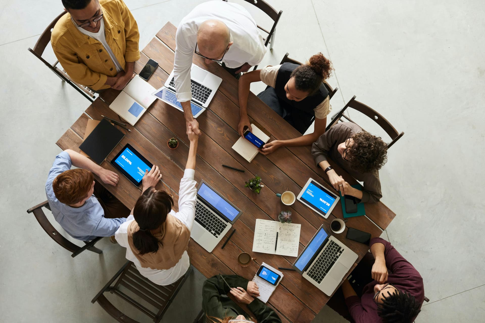 People sit around a wooden table, using laptops and smartphones, while two individuals shake hands in the center, suggesting a business meeting or collaboration in a well-lit, spacious room.