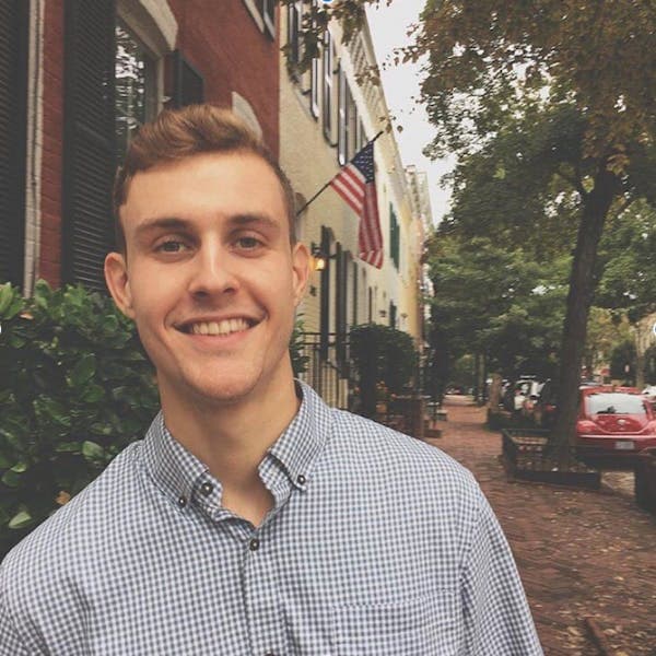 A young man smiles while standing on a brick sidewalk lined with trees, cars, and American flags, adjacent to a row of townhouses.