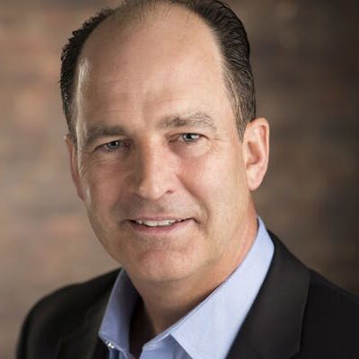 A middle-aged man with short hair is smiling slightly, dressed in a blue shirt and a dark blazer, standing against a blurred, neutral-tone background.