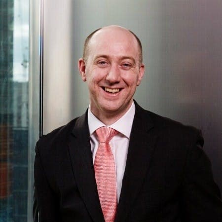 A man in a black suit with a pink tie is smiling, standing in front of a glass window with a blurred building background.