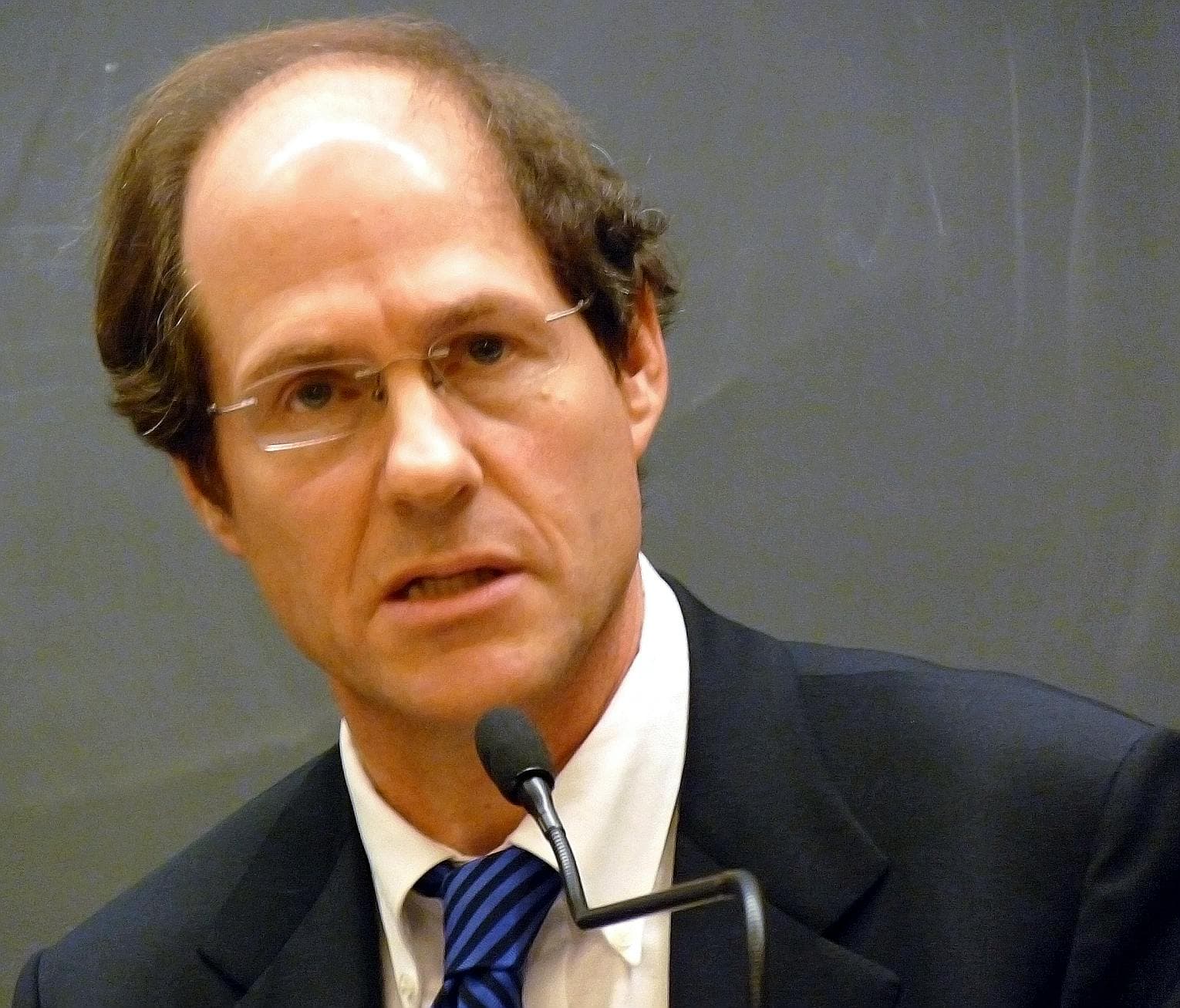 A man wearing glasses and a suit speaks into a microphone, against a plain, gray background.