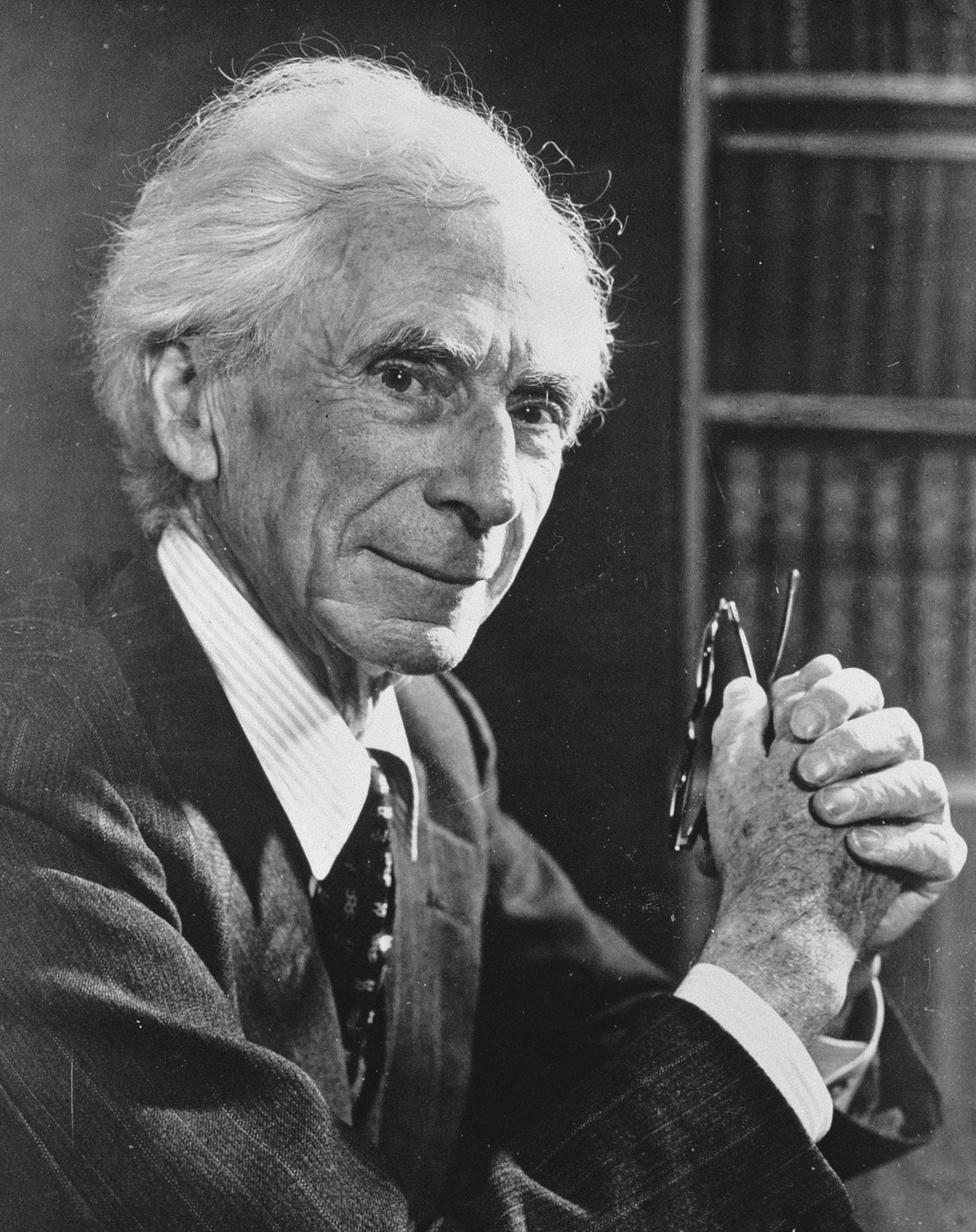 An elderly man with white hair clasps his hands, holding his glasses, while seated against a backdrop of bookshelves. He is wearing a dark suit and striped shirt.