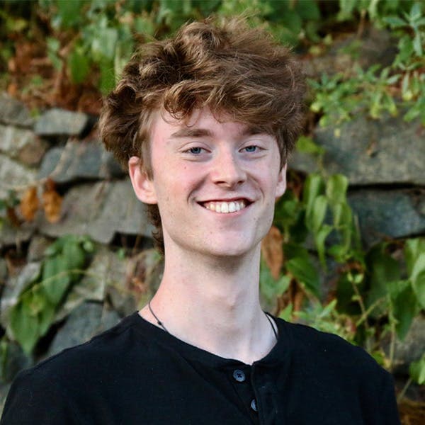 A person with tousled, light brown hair smiles while standing outdoors against a background of green foliage and stone.