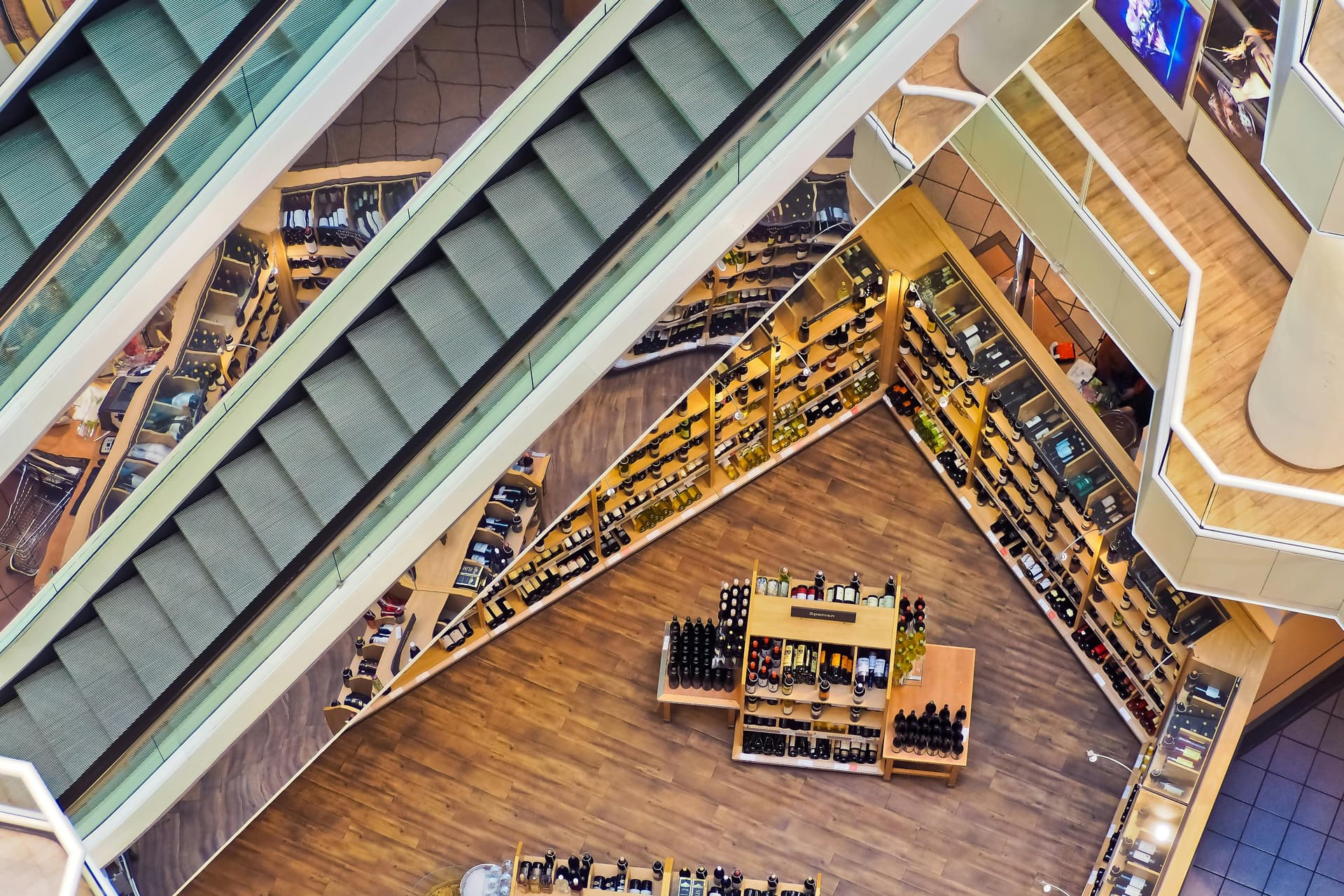 Escalators descending above a wine shop with wooden flooring, surrounded by shelves of wine bottles in a multi-level shopping mall.