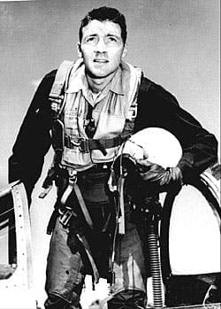 A black and white photo of a man standing on top of an open fighter plane. He holds a helmet in his left arm, while looking upward with a focused expression. He wears a full flight suit with visible straps and gear. The cockpit of the fighter plane is partially visible beside him.