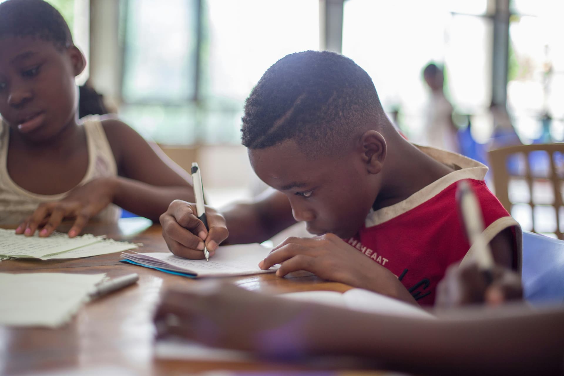 Child writes intently on paper at a table in a bright room, surrounded by other children engaged in similar activities.