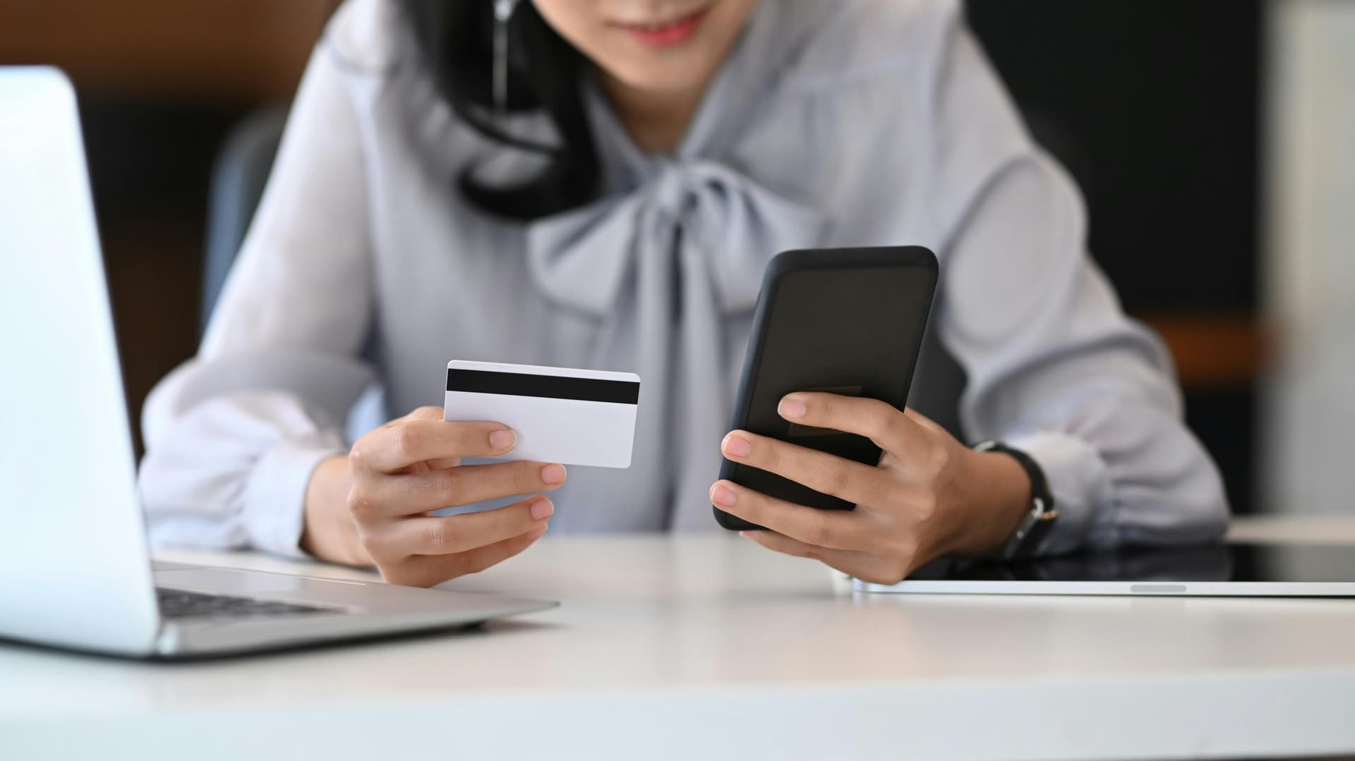 A person holding a credit card and a smartphone, making an online transaction at a desk with a laptop, in a modern indoor office setting.