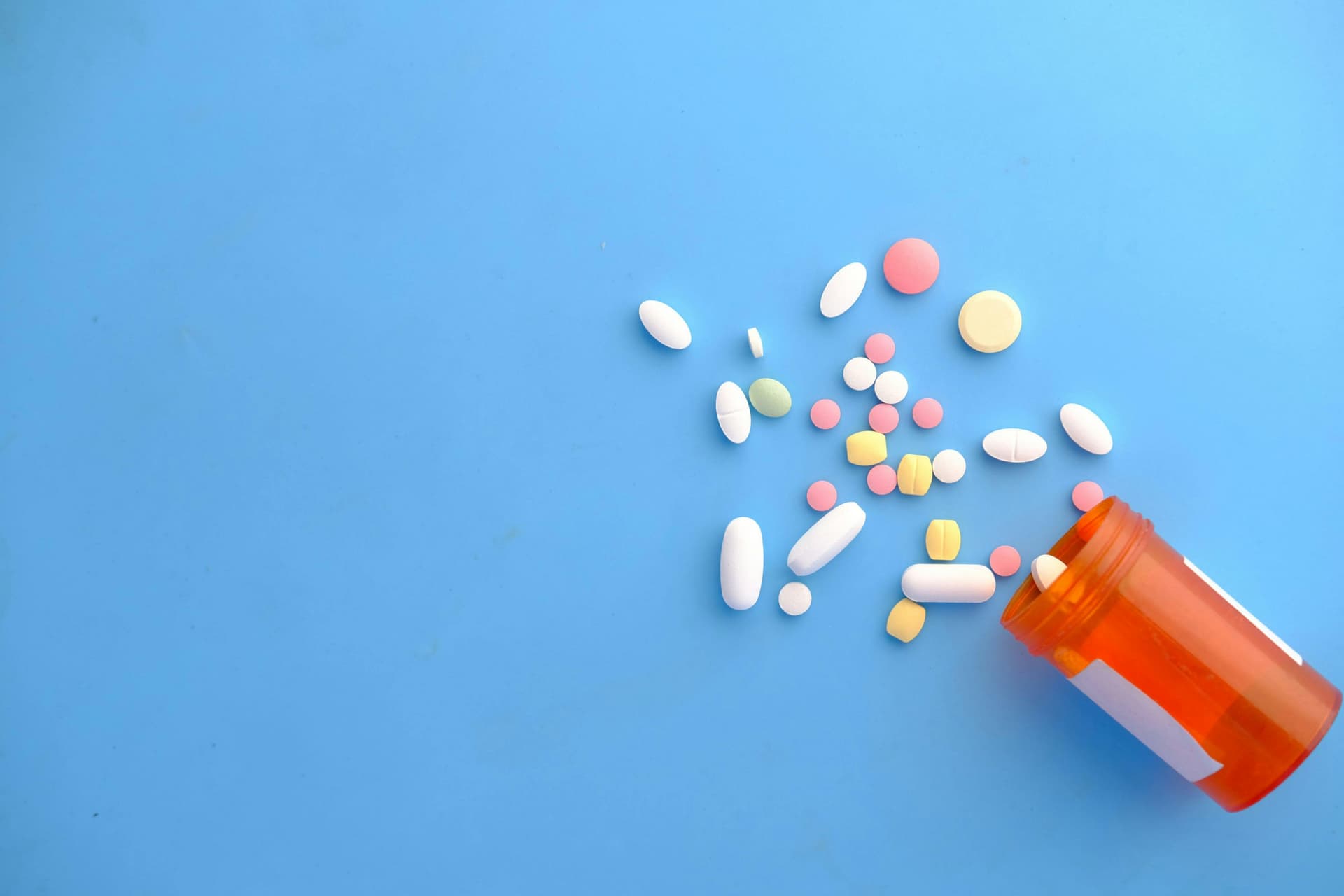 An orange prescription bottle spills variously shaped and colored pills across a blue surface, creating a scattered arrangement.