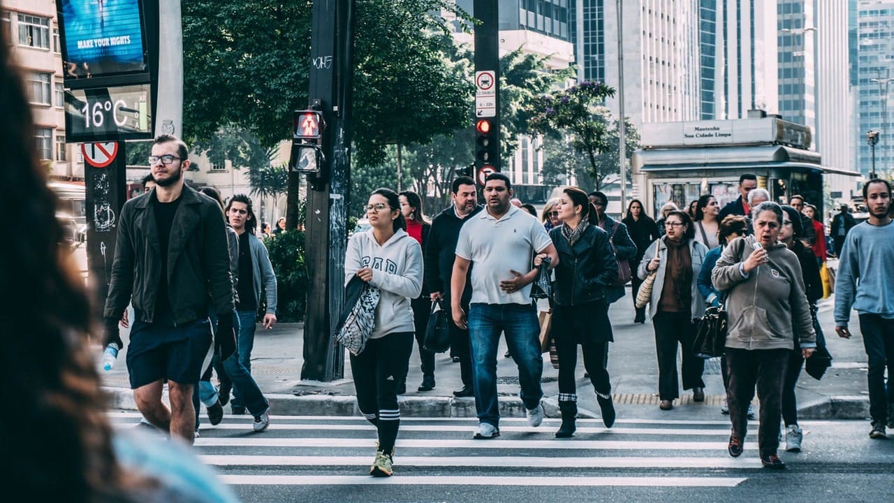 Pedestrians cross a city street at a crosswalk on a busy day, with tall buildings, trees, and a kiosk in the background. A digital sign displays "16°C MAKE YOUR RIGHTS."
