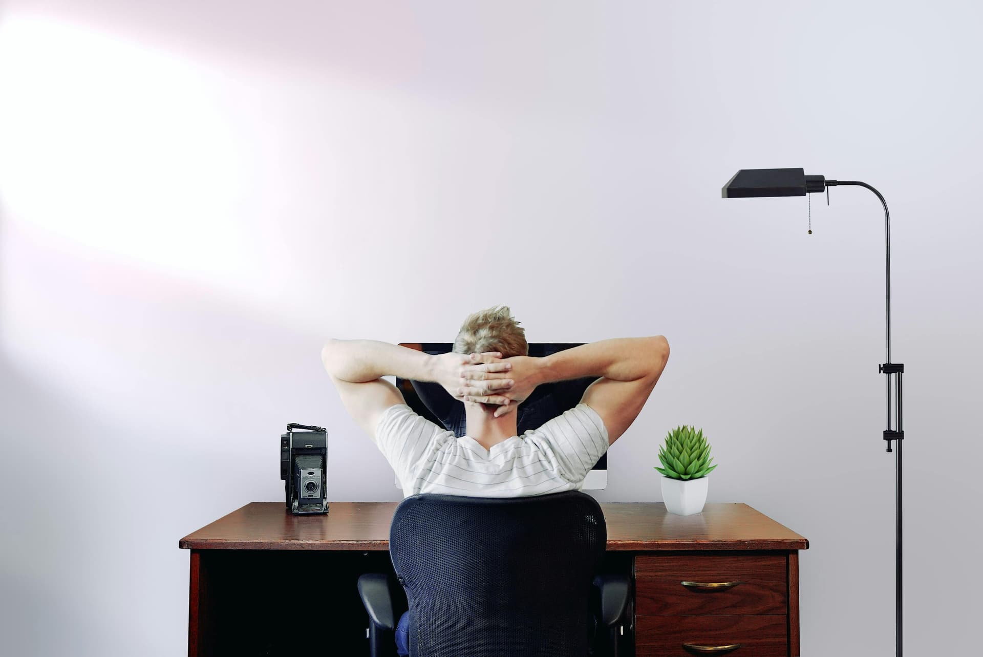 A person is leaning back in a chair with hands behind their head, facing a computer on a wooden desk in a minimalistic office with a plant and a floor lamp.