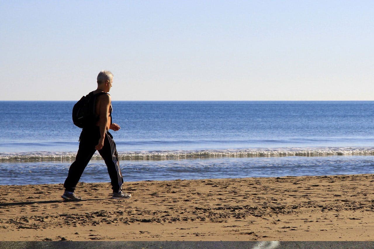 Person walking on sandy beach beside calm ocean, wearing black pants, tank top, and carrying a backpack, under a clear, blue sky.