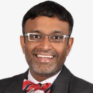 A person with short hair and glasses, smiling while wearing a black suit and red bow tie, against a plain white background.