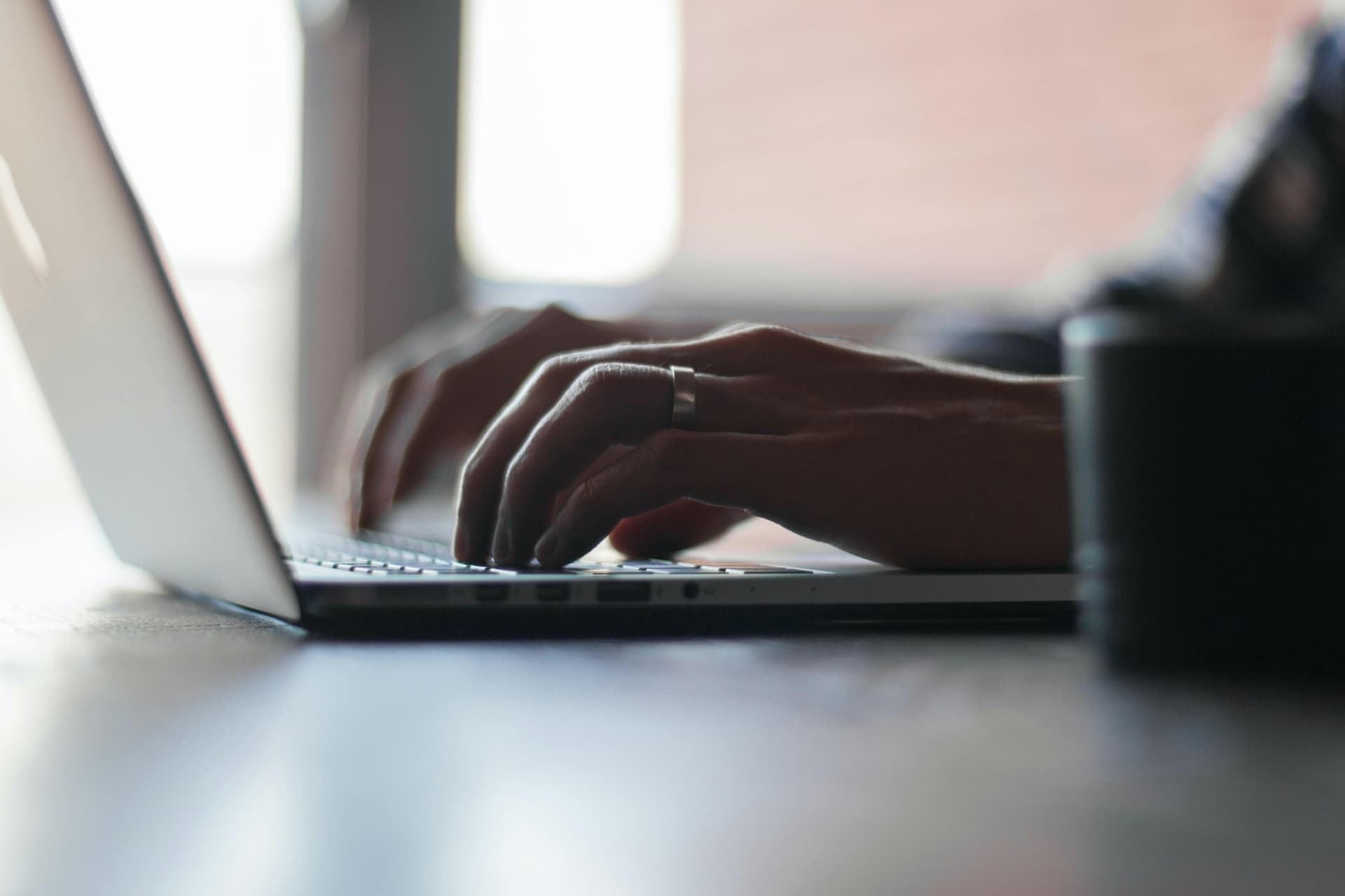 Hands typing on a laptop keyboard, a ring on one finger. In the background, light filters through a window, creating a softly lit workspace.