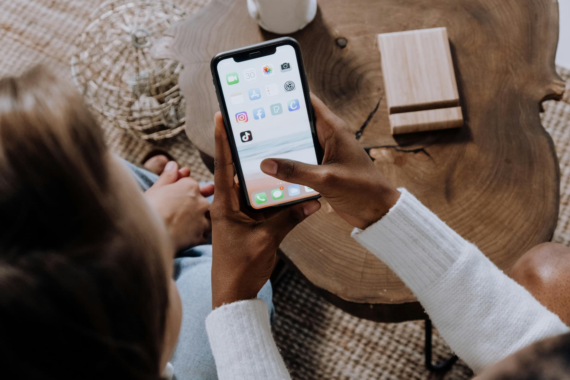 A person is holding a smartphone with several app icons visible, including messaging, shopping, and social media, while sitting beside another individual at a wooden table indoors.
