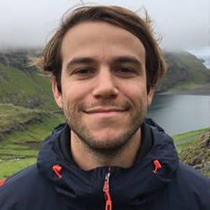 A man in a dark jacket smiles while standing outdoors, surrounded by green hills and a misty, overcast sky. A body of water and more hills are visible in the background.