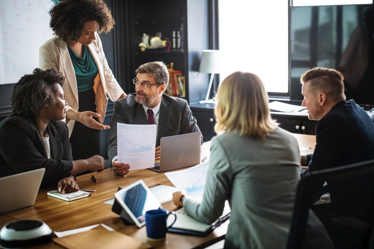 Business professionals collaborate at a conference table, discussing documents. One person is standing, gesturing towards seated colleagues in an office with laptops, notebooks, a whiteboard, and windows.