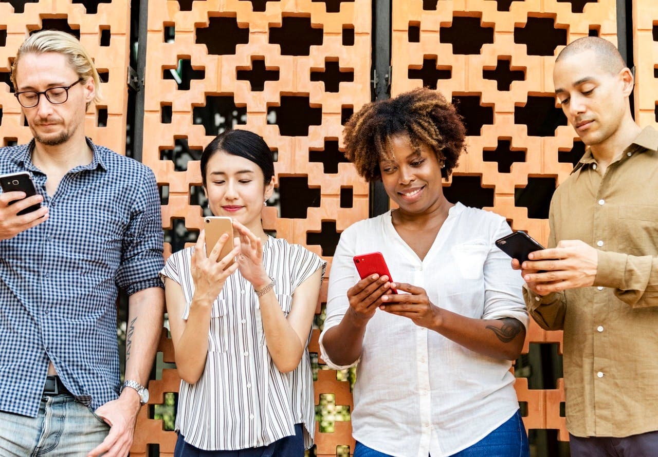 Four people stand against a patterned brick wall, all looking down and engaging with their smartphones. The context appears to be an outdoor setting with a decorative background.