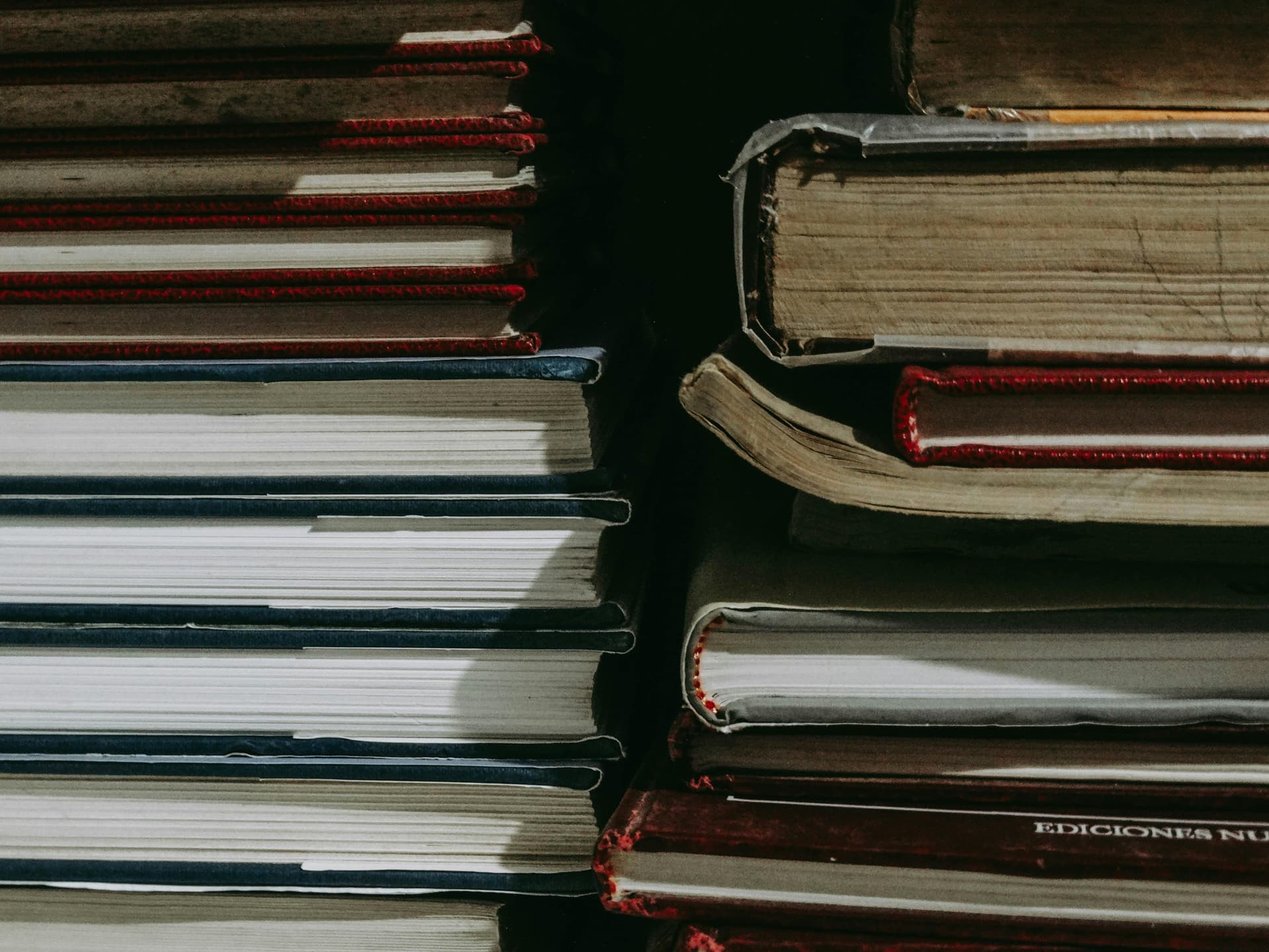 Stacks of old, worn books piled horizontally, their spines showing signs of age, placed closely together in a dimly lit space, suggesting an old library or a study.