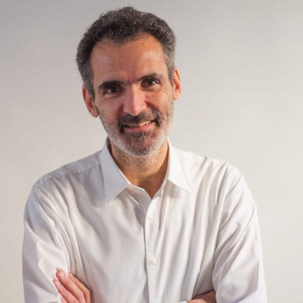 A man with graying hair and beard, wearing a white button-up shirt, stands with arms folded, smiling against a plain, light-colored background.