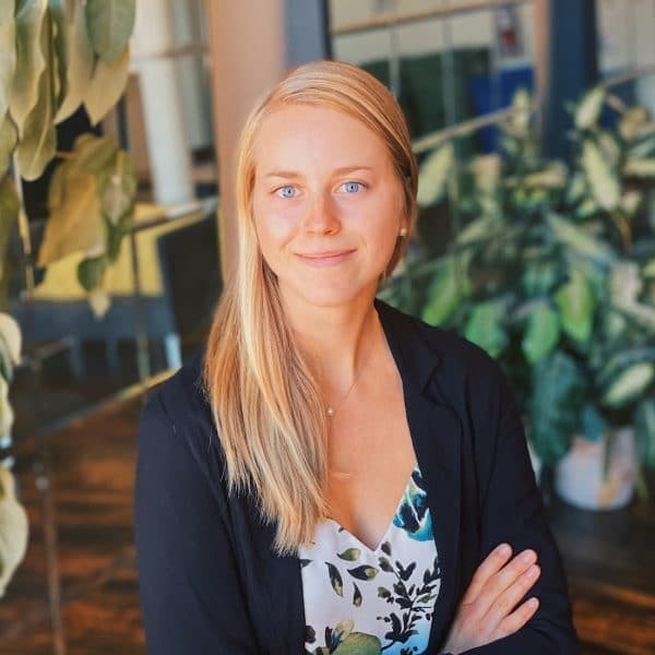 A woman with long blonde hair stands confidently with folded arms, wearing a floral top and black blazer, in an indoor setting decorated with large green plants and modern furnishings.