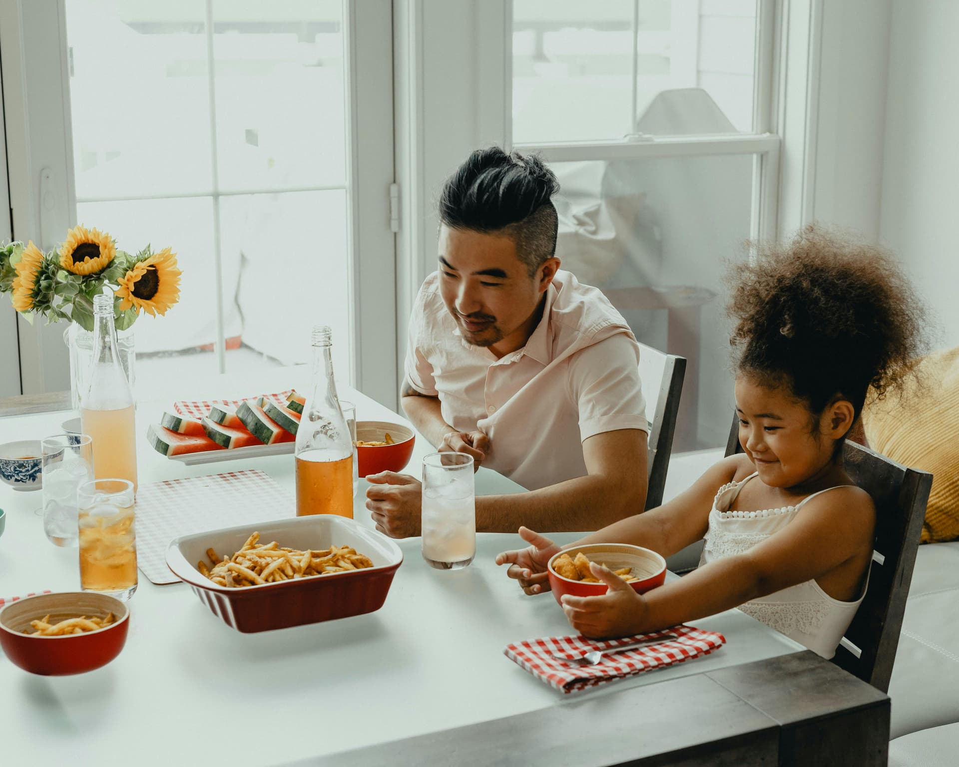 A man and a girl sharing a meal at a table with sunflowers, fries, watermelon, and drinks in a bright, sunlit dining area.