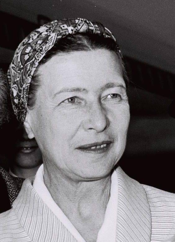 A black and white headshot of a woman wearing a patterned headscarf. She looks slightly to the side with a subtle smile. She wears a light-colored, collared coat in an indoor setting with dim lighting.