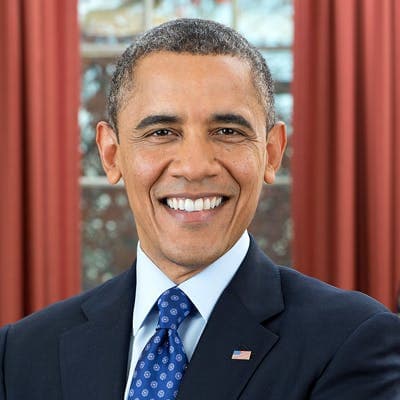 A man in a suit and blue patterned tie smiles broadly, standing in front of red curtains with a window in the background.