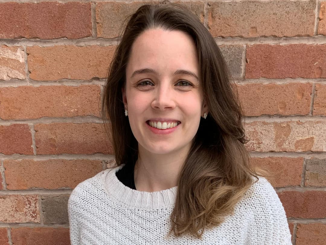 A woman with long brown hair and a white knit sweater is smiling in front of a red brick wall background.