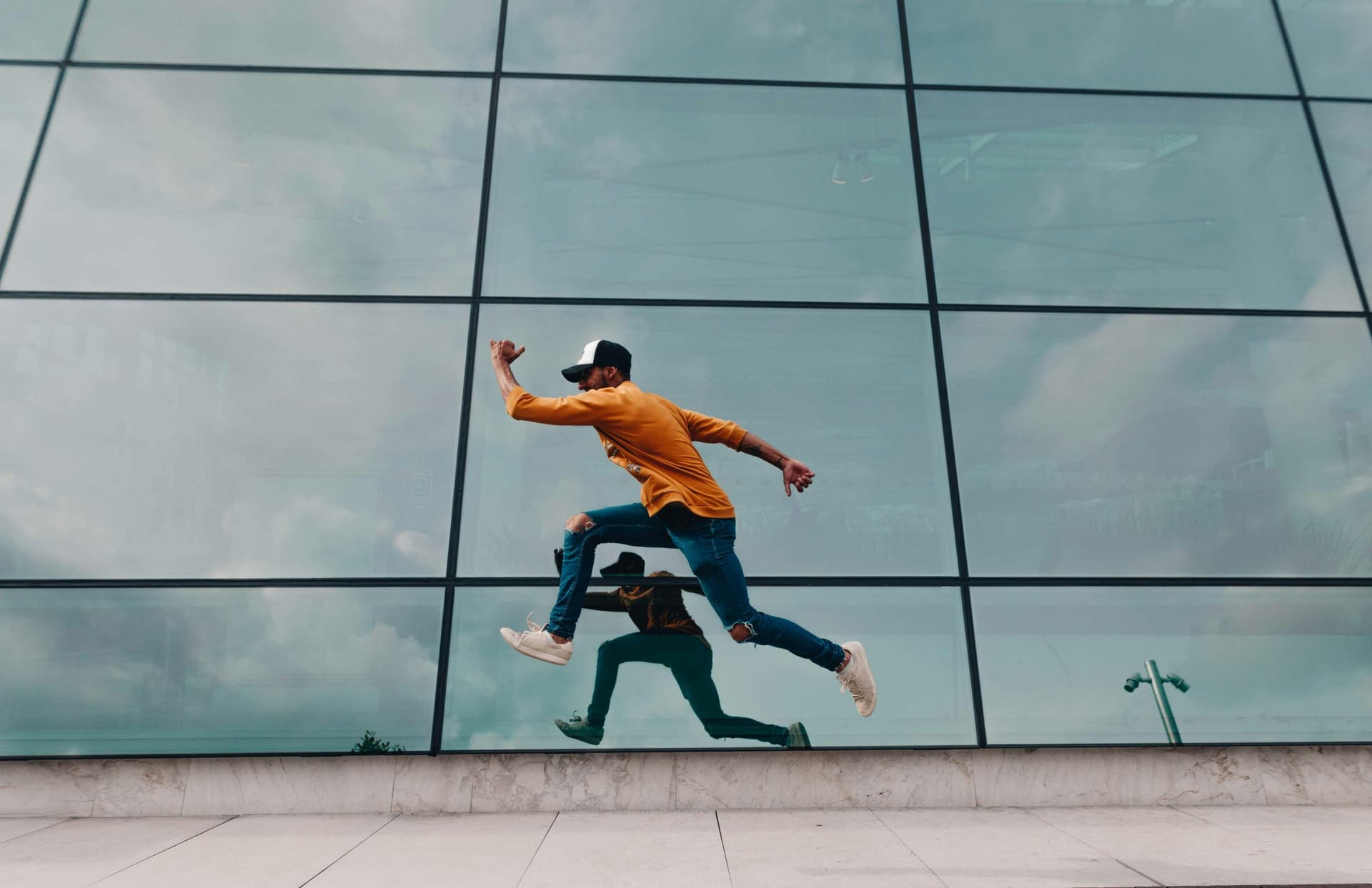 A person in a yellow sweater and cap leaps mid-air, reflected in a large glass building facade, against a cloudy sky backdrop.