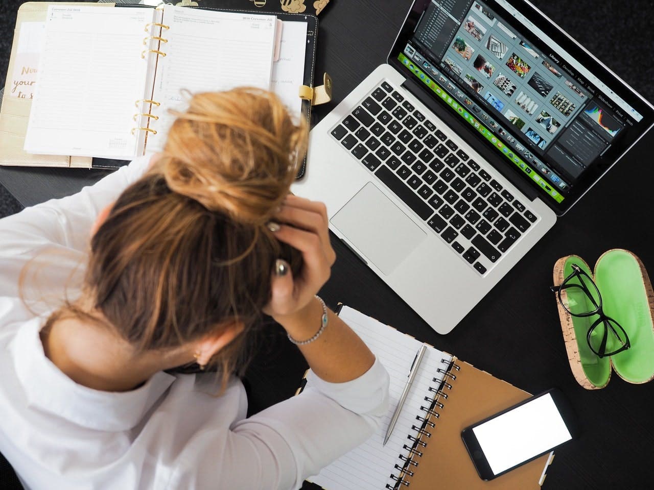 Person with head in hands, surrounded by open notebook, laptop, smartphone, and glasses case on a cluttered desk. Laptop screen displays multiple thumbnail images.
