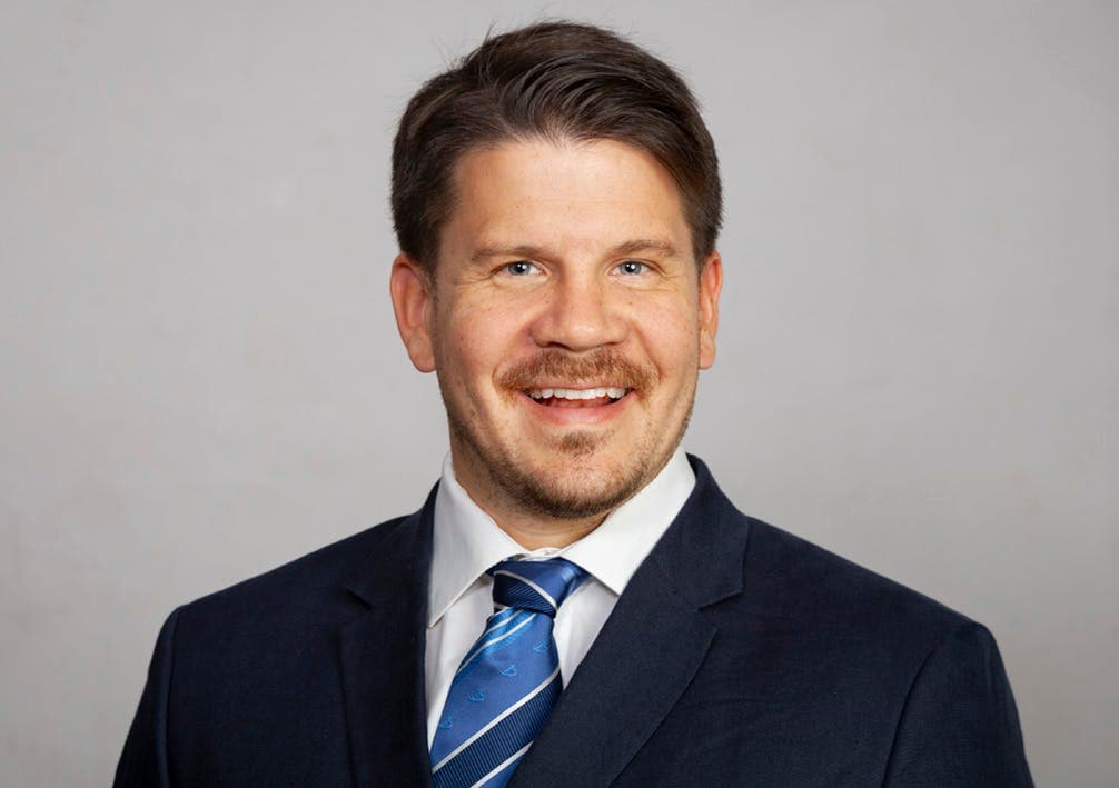 The image is a professional headshot of a smiling man wearing a dark suit, white shirt, and blue striped tie. He has short brown hair and a neatly trimmed beard, set against a plain background.