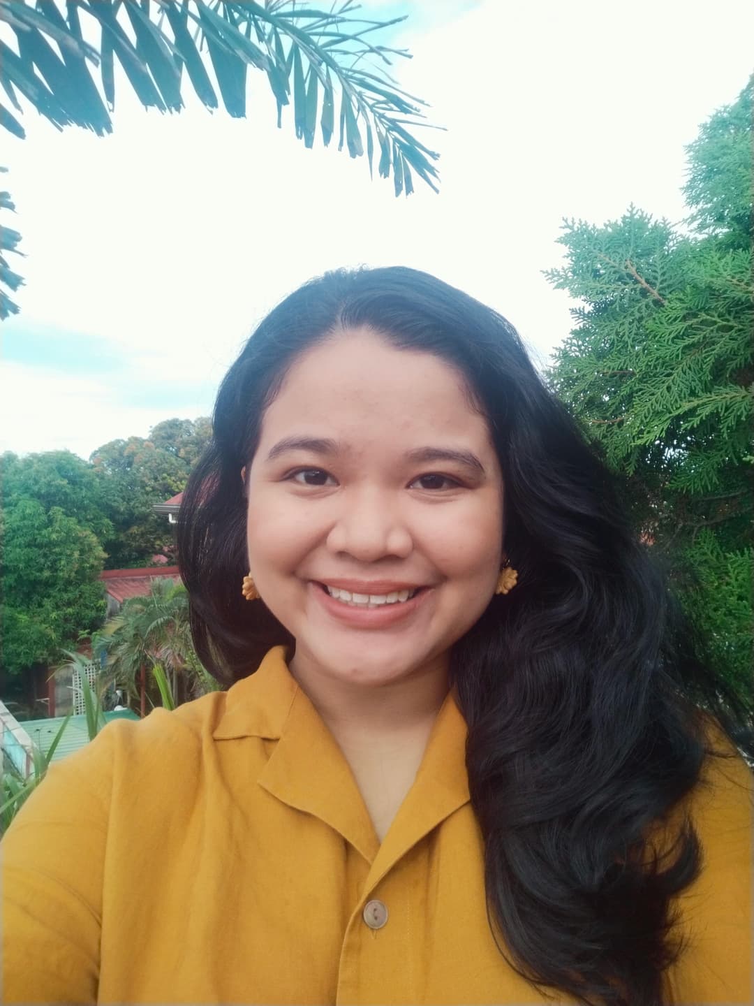 Woman smiling outdoors, wearing a mustard yellow shirt and flower earrings. She is surrounded by green trees and plants, under a clear sky.