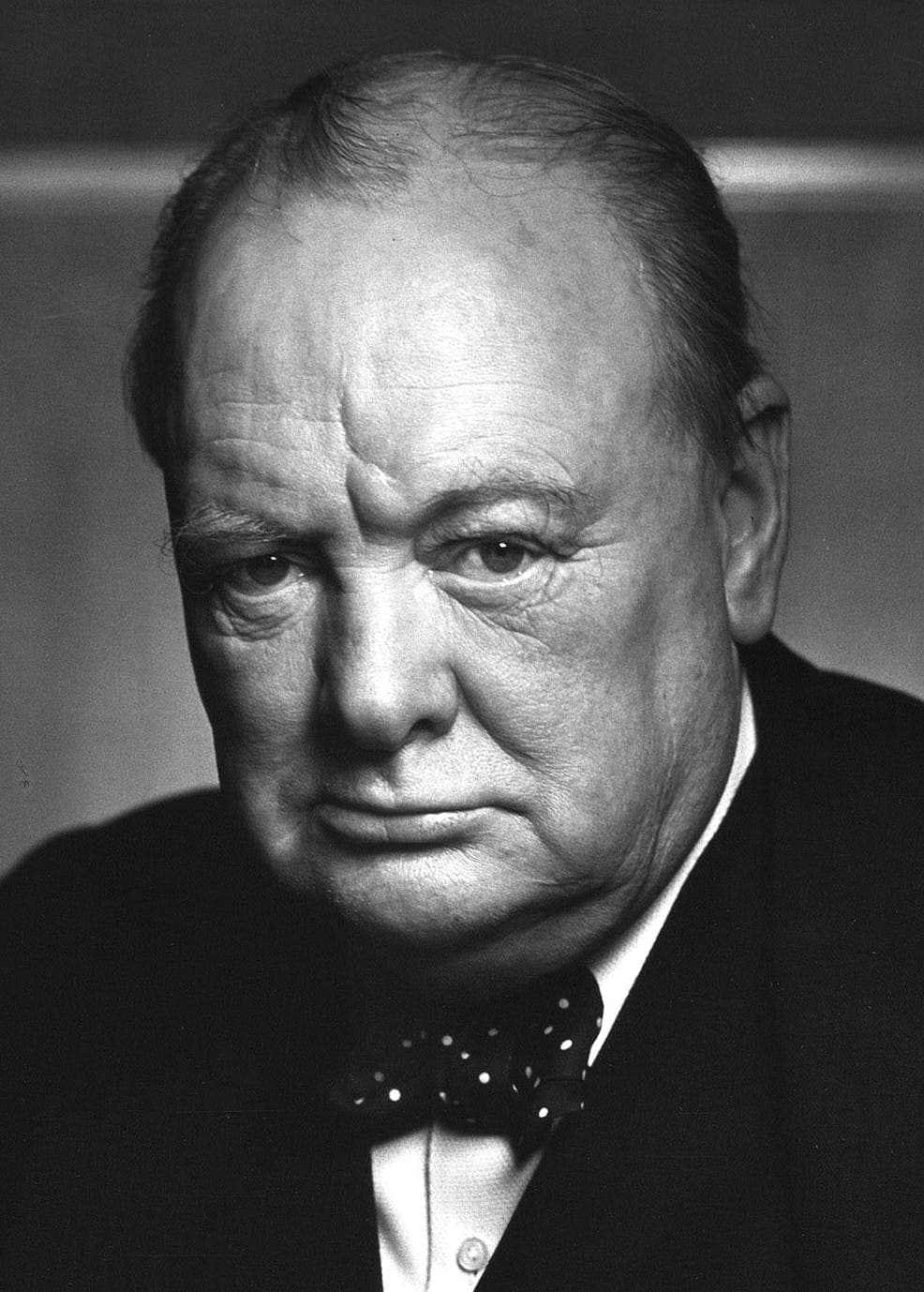 Older man wearing a polka-dotted bow tie and dark suit, gazing sternly at the camera against a neutral background.