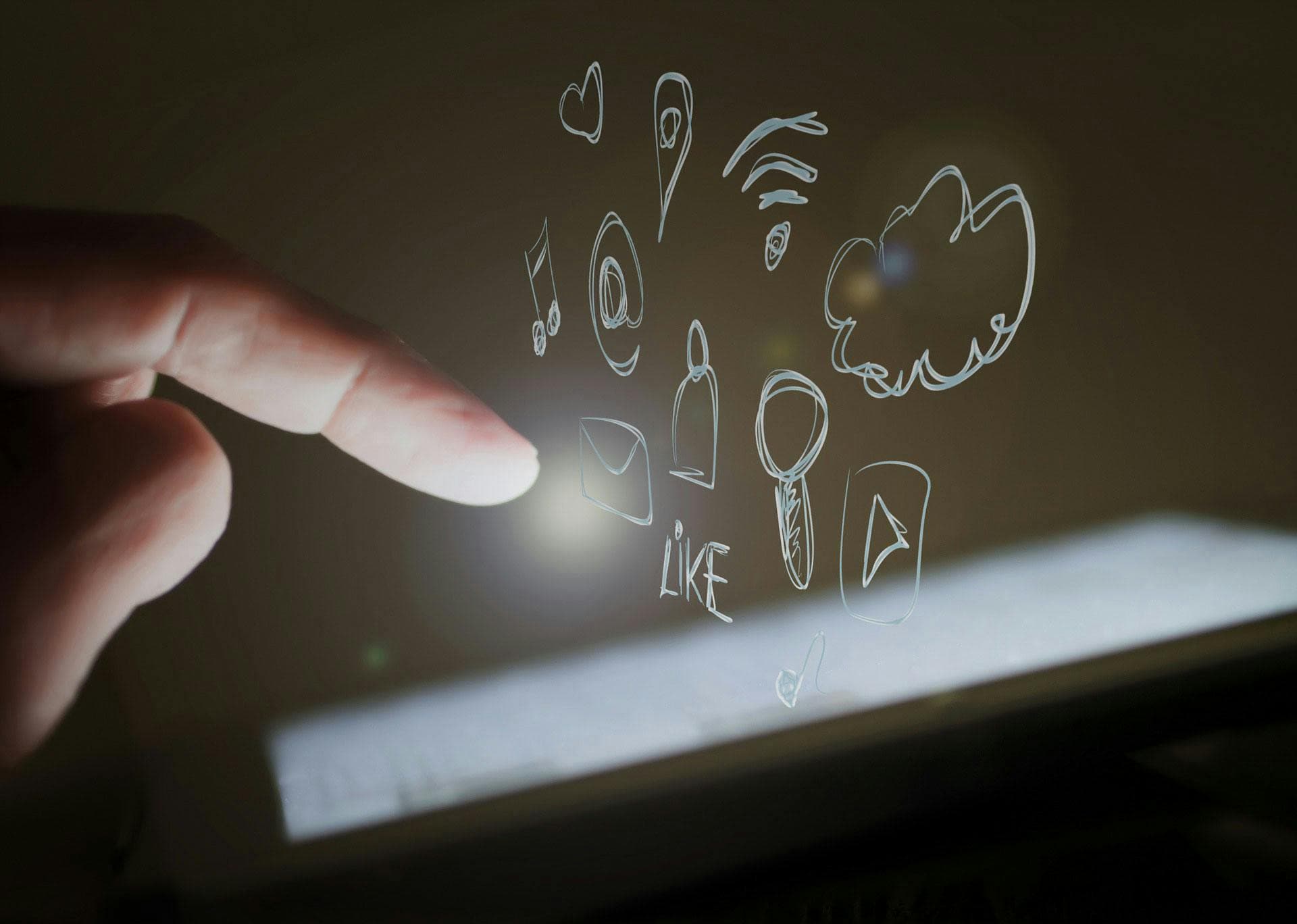 A finger points toward a tablet screen, illuminating various holographic app icons and symbols, including a heart, Wi-Fi signal, music note, and the word "LIKE," in a dark environment.