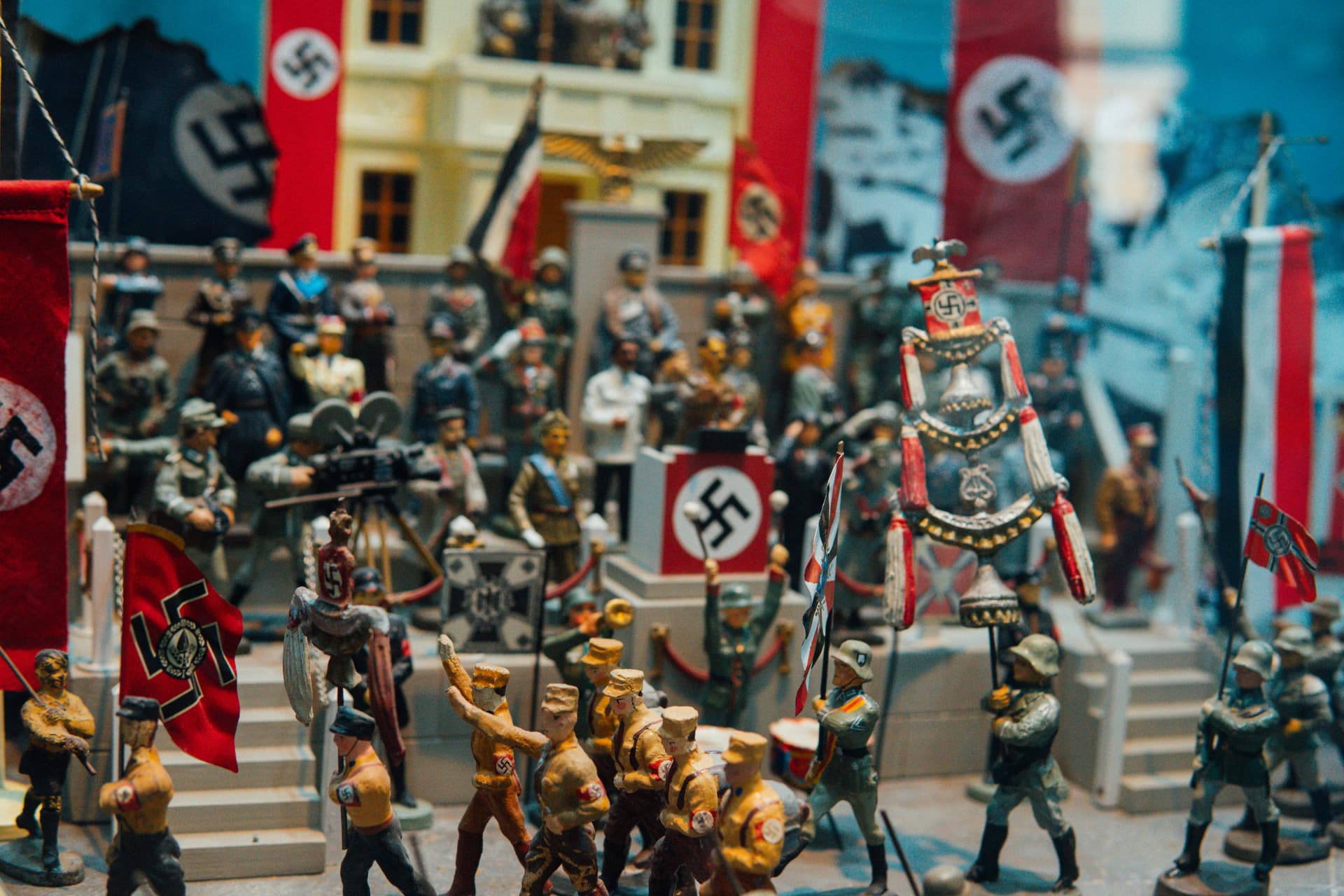 The image depicts a detailed diorama or model display of a Nazi rally, with miniature figures of Nazi soldiers, officials, and flags prominently featuring swastikas. The scene is filled with figures arranged in a setting that likely represents a propaganda event or military parade during the Third Reich. The flags and symbols of Nazi Germany are clearly visible throughout the scene, conveying the historical context of the time.