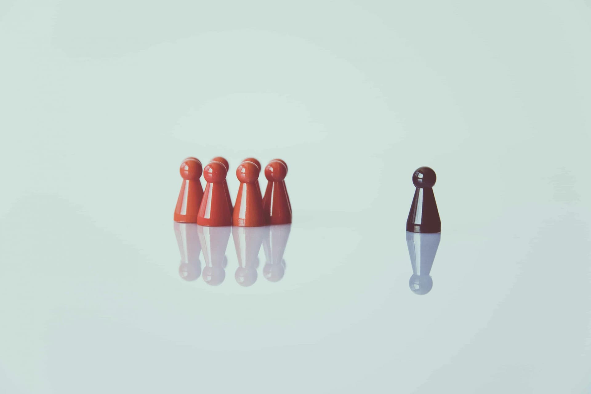 A single black pawn stands apart from a group of five red pawns on a reflective, white surface.