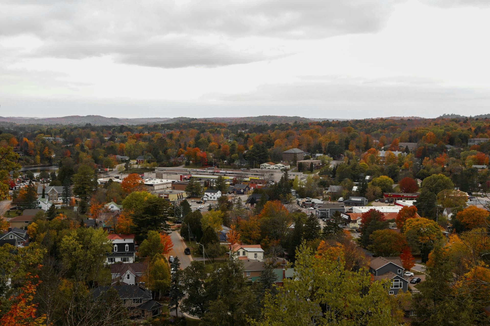 A panoramic view of a small town surrounded by autumn foliage, with vibrant red, orange, and yellow trees scattered among houses and buildings under a cloudy sky.