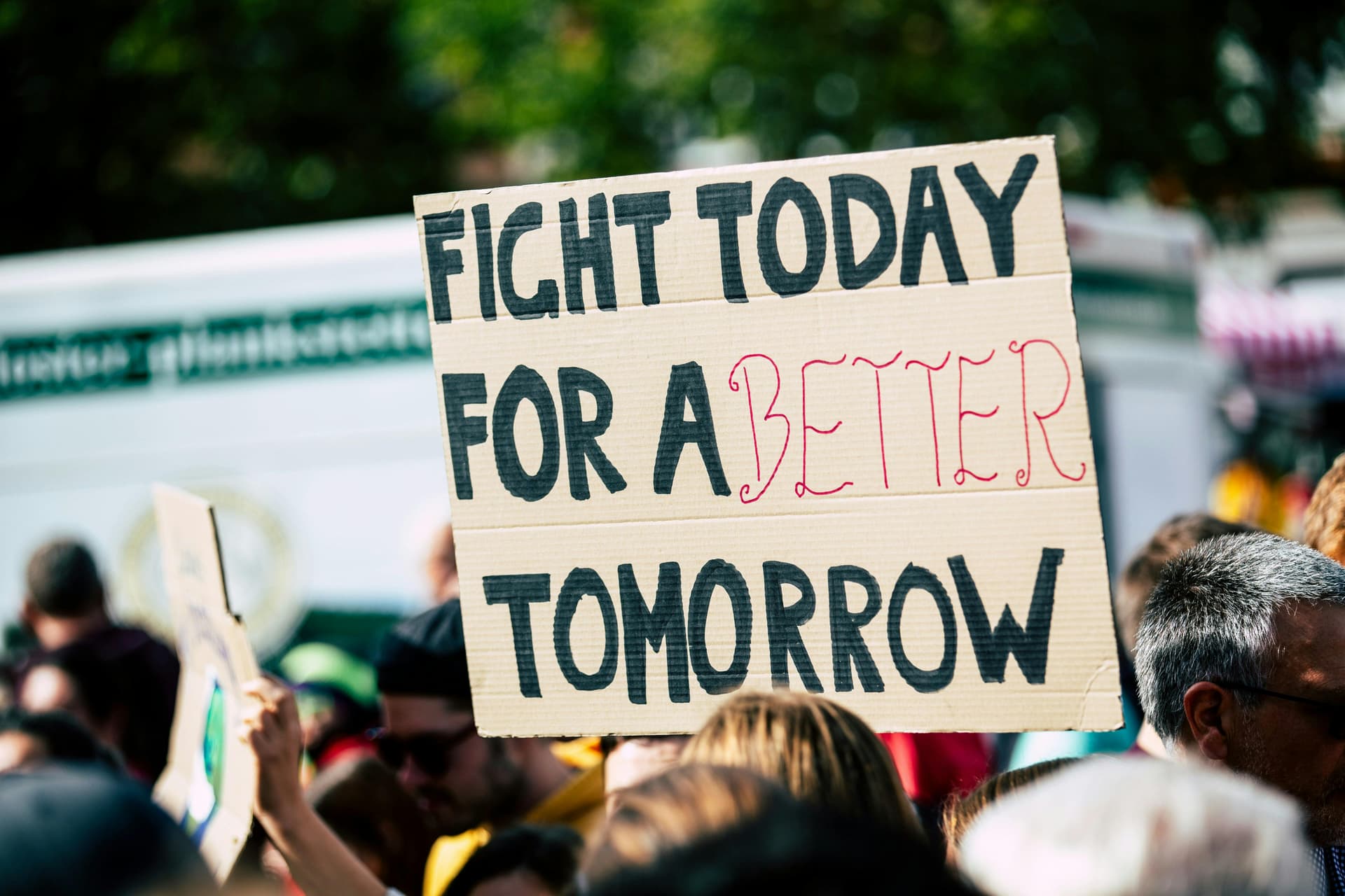 A close-up of a protest sign held in a crowd. The sign, made from cardboard, reads, "FIGHT TODAY FOR A BETTER TOMORROW" in bold black letters, with the word "BETTER" highlighted in red. The background shows blurred people and trees, suggesting an outdoor event.