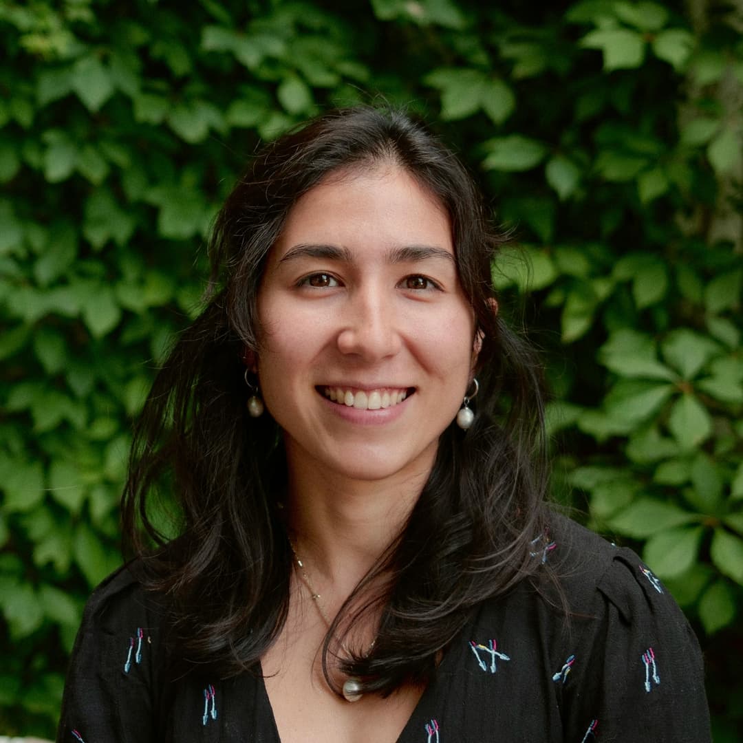 A woman with mid-length hair wearing pearl earrings and necklace stands behind a green foliage.