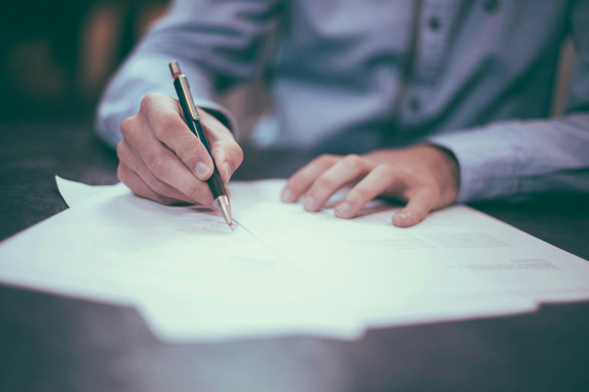 A person writing on papers with a pen, likely engaged in some form of documentation or signing a contract. The individual is wearing a light-colored shirt, and the focus is on the hands and the pen. The scene suggests work or professional documentation in progress.